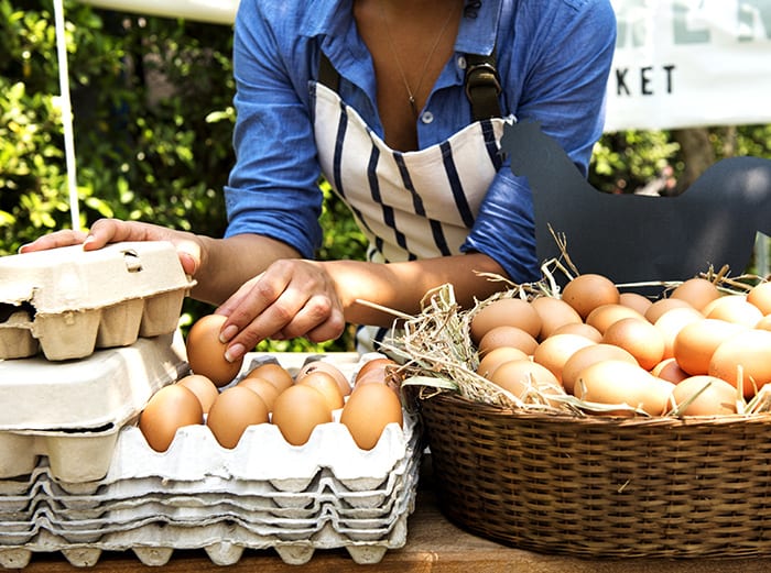 eggs in baskets and crates set out on table, most are brown eggs with some white eggs. A woman hovers above the eggs as she places them into the crates and baskets. She is wearing a long sleeve blue shite with a a blue striped apron.